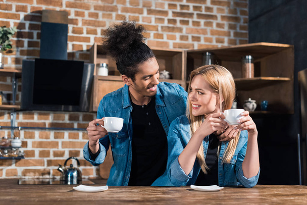 portrait of smiling multiracial young couple with cups of coffee in kitchen at home - Foto, Imagen
