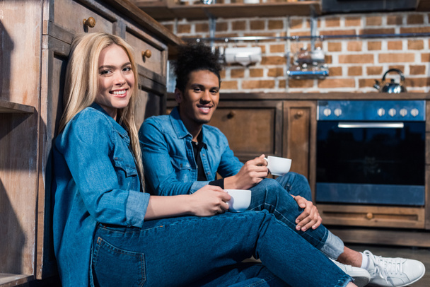 uśmiechający się międzyrasowy pary młodych z filiżanki kawy, patrząc na kamery w kuchni w domu - Zdjęcie, obraz