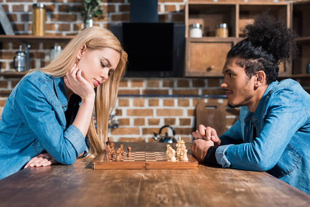 side view of multiethnic young couple playing chess together in kitchen - Foto, afbeelding