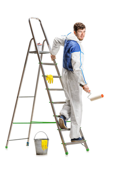 Young male decorator painting with a paint roller climbed a ladder isolated on white background. - Photo, image