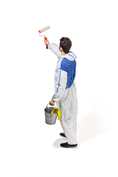 Young male decorator painting with a paint roller climbed a ladder isolated on white background. - Фото, зображення