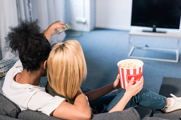 back view of multiracial couple with popcorn watching film together at home - 写真・画像