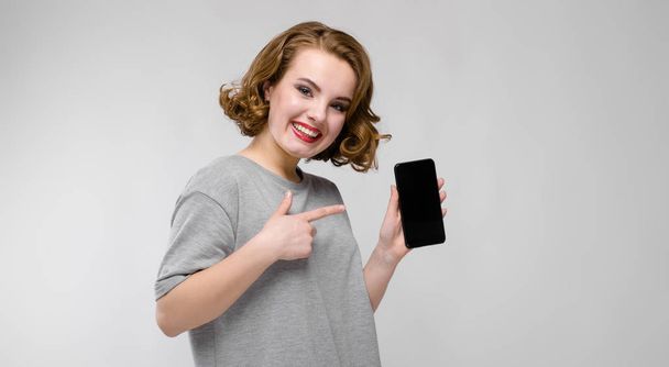 Gyönyörű vörös hajú boldog fiatal nő, telefon, üres képernyő a szürke háttér portréja - Fotó, kép