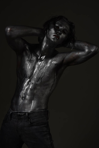 Guy posing with confident sexy expression. Fashion and erotic concept. Man with nude torso covered with shimmering silver paint, dark background. Macho with streams of sweat or paint on naked chest. - Photo, image