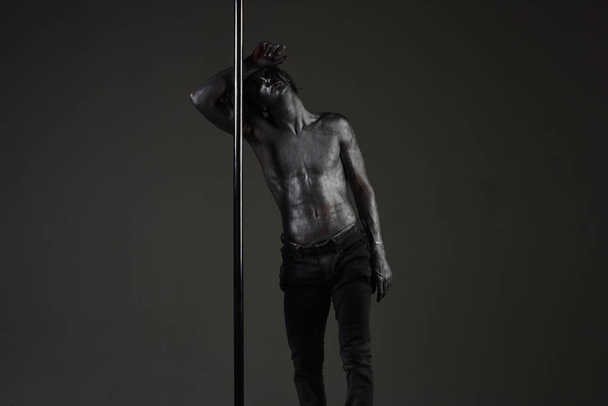 Tired dancer concept. Guy lean on metallic pole. Man with nude torso covered with shimmering silver paint, dark background. Man with naked chest, athlete, sportsman performing pole dancing moves. - Foto, Imagem