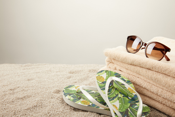 close up view of stack of towels, sunglasses and summer flip flops on sand on grey backdrop - Photo, Image
