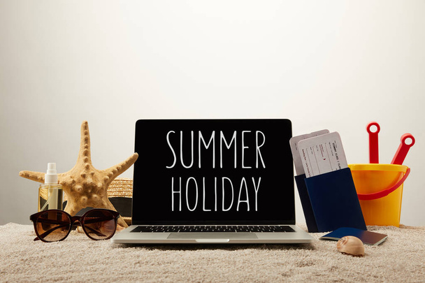 vista de cerca de la computadora portátil con letras de vacaciones de verano, estrella del mar, gafas de sol, pasaportes con entradas y cubo de juguete en la arena sobre fondo gris
 - Foto, Imagen