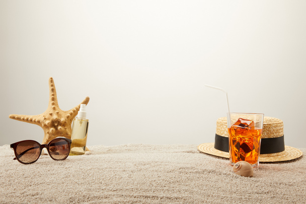 close up view of cocktail with ice, straw hat, sunglasses and tanning oil on sand on grey backdrop - Photo, Image