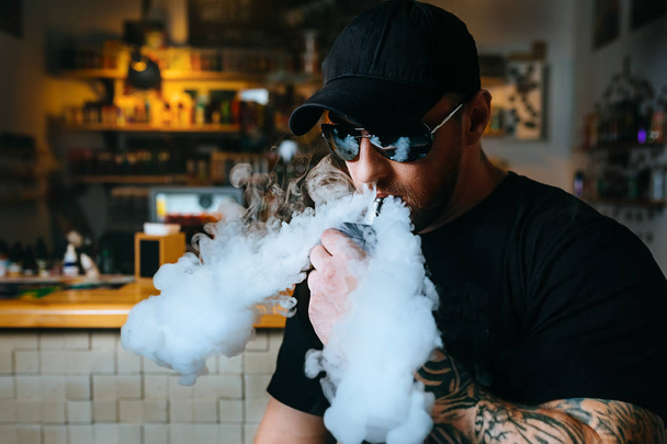 Βάναυση γενειοφόρος άνδρας με γυαλιά ηλίου καπνίζει ένα ηλεκτρονικό τσιγάρο και απελευθερώνει σύννεφα ατμού στο κατάστημα vape. - Φωτογραφία, εικόνα