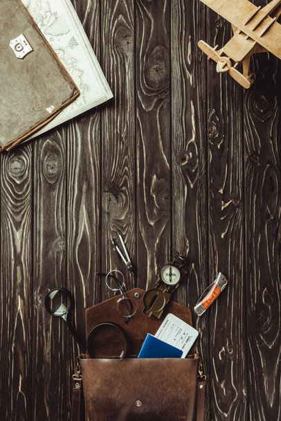 Ansicht der Anordnung von Holzspielzeug-Flugzeug, Tasche mit Lupe, Taschenmesser, Reisepass, Ticket, Brille und Kompass auf dunkler Holztischplatte - Foto, Bild