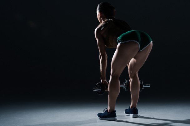 back view of sexy female bodybuilder lifting dumbbells, on black    - Фото, изображение