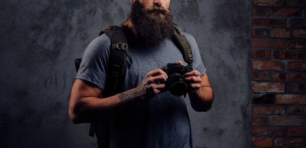 Портрет красивого бородатого путешественника в шляпе с рюкзаком и татуированными руками, держит фотокамеру, изолированную на темном фоне
. - Фото, изображение