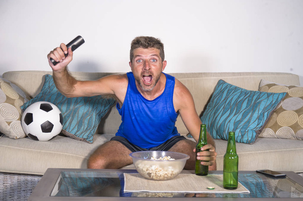 young attractive man happy and excited watching football match on TV celebrating victory goal crazy and spastic with beer popcorn and soccer ball  - Foto, Bild