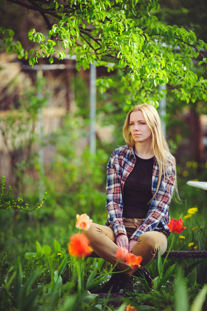 мила блондинка сидить в домашньому саду, на фоні багатьох зелених дерев і тюльпанів, красивий портрет сільського стилю, простий домашній образ
 - Фото, зображення