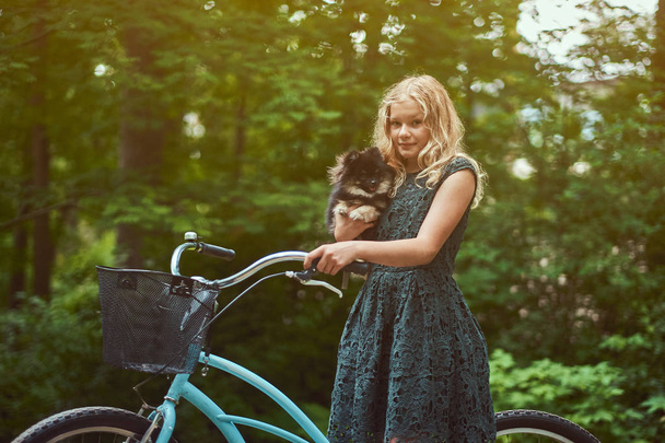 Porträt eines kleinen blonden Mädchens in einem lässigen Kleid, hält niedlichen Spitz-Hund, in einem Park. - Foto, Bild