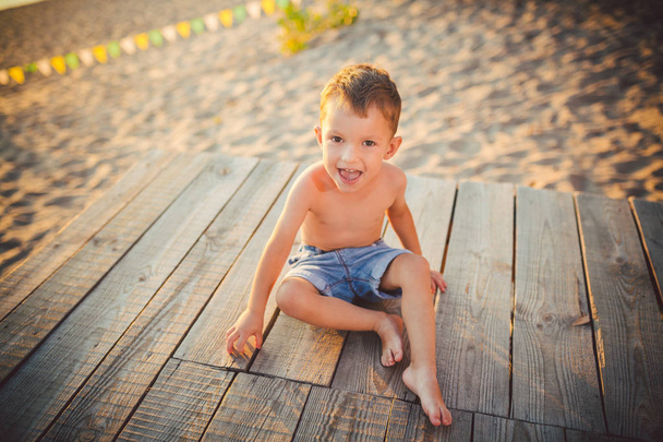 The theme is child and summer beach vacation. A small Caucasian boy sits sideways on a wooden pier and admires the view of the sandy beach and the pond, the river. With bare legs in blue denim shorts - Photo, Image