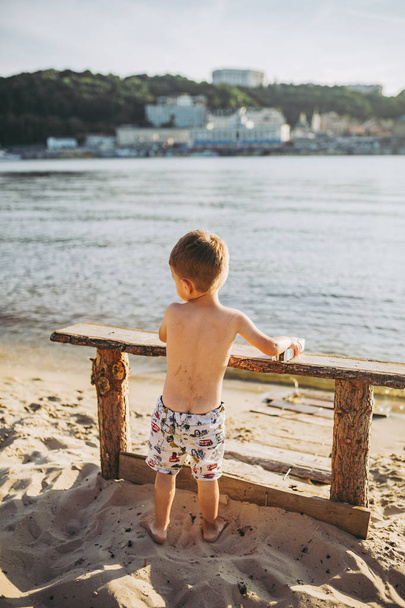 El tema es un niño y vacaciones de verano en la playa. Un pequeño chico caucásico sube a un banco de madera en la orilla del río en un soleado día de verano en una playa de arena con vistas a la ciudad
 - Foto, imagen