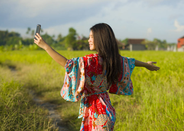 young beautiful and happy Asian Chinese tourist woman on her 20s with colorful dress taking selfie pic with mobile phone camera on tropical field - 写真・画像