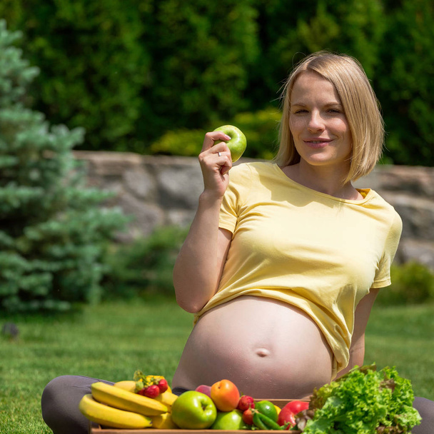 Pregnant woman sitting on grass and holding an apple in hand - Foto, Imagen