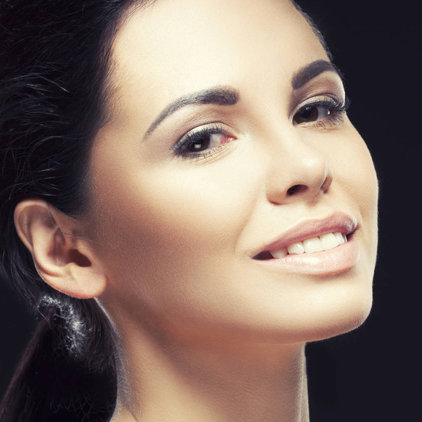 Portrait of young brunette woman with natural makeup and tender smile on face against black background - Photo, Image