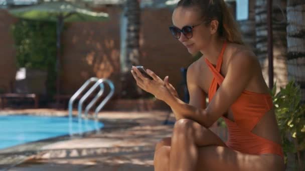 blond girl with ponytail in swimsuit sits in shady place near swimming pool and texts sms on smartphone - Кадри, відео