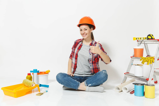 Γυναίκα στο πορτοκαλί προστατευτικό κράνος που κάθεται στο πάτωμα με τα όργανα για δωμάτιο διαμέρισμα ανακαίνιση που απομονώνονται σε λευκό φόντο. Ταπετσαρία, αξεσουάρ για την κόλληση, εργαλεία ζωγραφικής. Επισκευή concept home. - Φωτογραφία, εικόνα