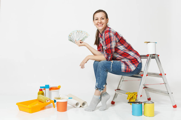 Woman holds bundle of dollars, cash money, sits on ladder with instruments for renovation apartment isolated on white background. Wallpaper accessories for gluing painting tools. Repair home concept. - Photo, Image
