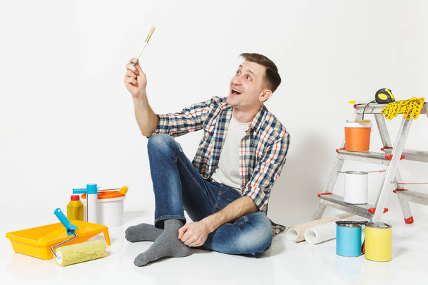 Smiling fun happy man sitting on floor with paint brush, instruments for renovation apartment room isolated on white background. Wallpaper, gluing accessories, painting tools. Repair home concept. - Photo, Image
