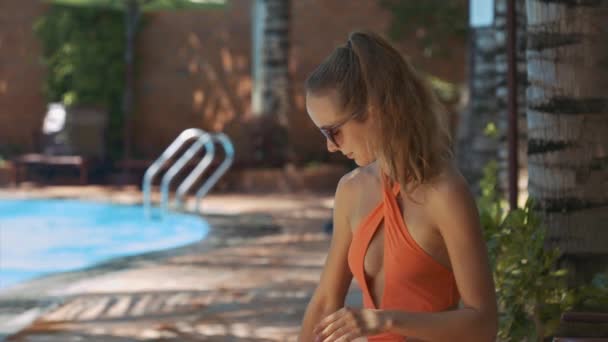closeup slim blond girl in orange bikini smears arms with sunscreen sitting in shade on sunny day - Materiał filmowy, wideo