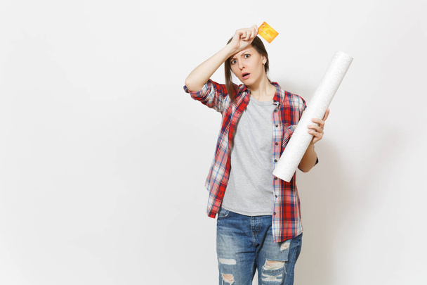 Young exhausted woman in casual clothes holding credit card and wallpaper roll and keeping hands near face isolated on white background. Instruments for renovation apartment room. Repair home concept. - Φωτογραφία, εικόνα