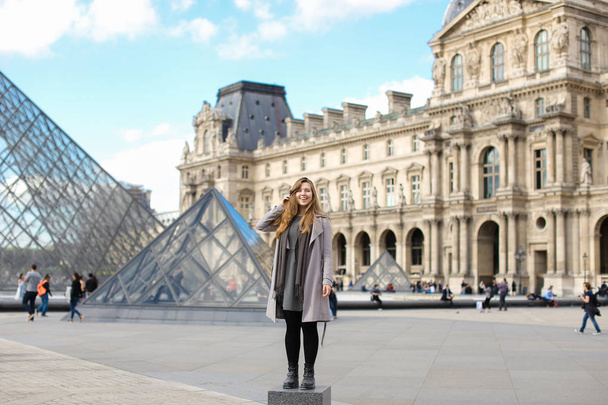 Όμορφη γυναίκα, φορώντας γκρι παλτό και στέκεται κοντά στο Λούβρο και γυάλινη πυραμίδα στο Παρίσι. - Φωτογραφία, εικόνα