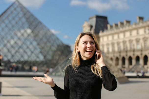 Young woman standing near Louvre and glass pyramind in black dress in Paris, talking by smartphone. - Фото, зображення