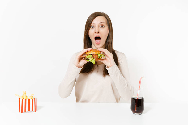 Beautiful young woman sitting at table with burger, french fries, cola in glass bottle isolated on white background. Proper nutrition or American classic fast food. Advertising area with copy space. - Photo, Image