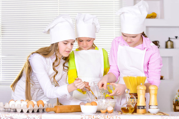 Cute little girls in chef's hats and aprons preparing dough in the kitchen at home - Photo, image