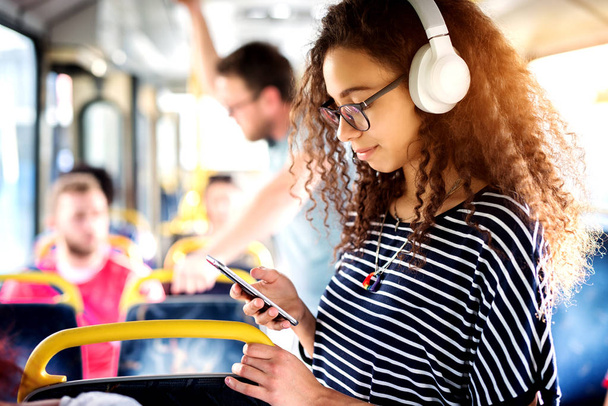 молодая веселая женщина в наушниках, стоящая в автобусе и пользующаяся мобильным телефоном
 - Фото, изображение