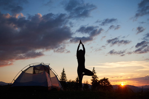 Vue arrière de la silhouette jeune femme debout sur la jambe dans un arbre de yoga pose sur le sommet de la colline près de la tente à l'aube sous le ciel du soir avec des paysages parfaits sapins, montagnes et coucher de soleil
 - Photo, image