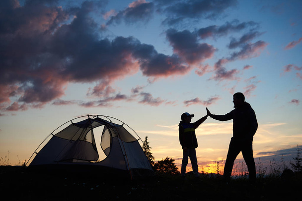 男と子のハイカーのシルエットがお互いに背景に山とそれらの上に昇る太陽の夜明けのテント近く高 5 を与えます。健康的なライフ スタイルのコンセプト. - 写真・画像