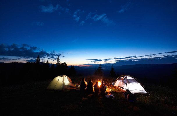 Молодые семейные туристы мать, отец, два сына отдыхают в палатках в горах, сидят на бревне у костра и две освещенные палатки, наслаждаясь прекрасным видом на вечернее небо, закат, звезды
 - Фото, изображение