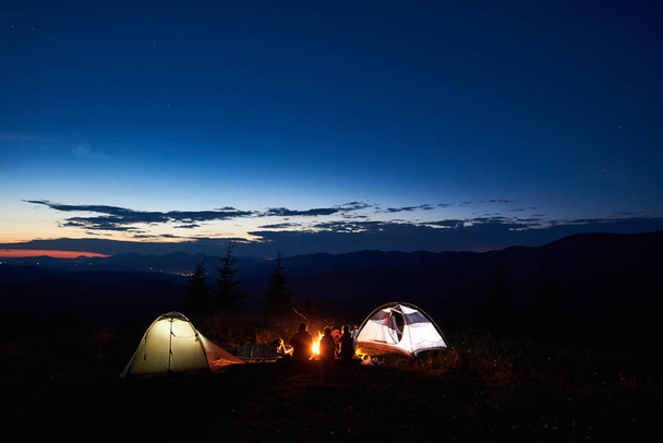 Jóvenes turistas familiares madre, padre, dos niños que se relajan al acampar en las montañas, sentados en el tronco junto a la fogata y dos tiendas de campaña iluminadas, disfrutando de una vista increíble del cielo nocturno, puesta de sol, estrellas
 - Foto, imagen