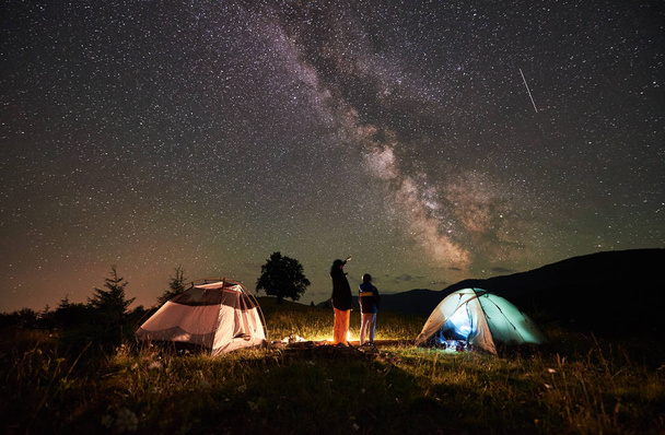 Voltar vista mãe e filho turistas descansando no acampamento em montanhas, de pé ao lado da fogueira e duas tendas, olhando para o céu noturno cheio de estrelas e Via Láctea, desfrutando de cena noturna. Mulher apontando para o céu
 - Foto, Imagem