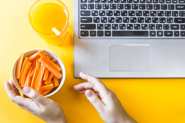 Eine Frau nimmt ihre Hände mit Karotten und einem Glas Saft, das neben dem Laptop steht. Pause von der Arbeit für einen Snack. gesundes Ernährungskonzept - Foto, Bild
