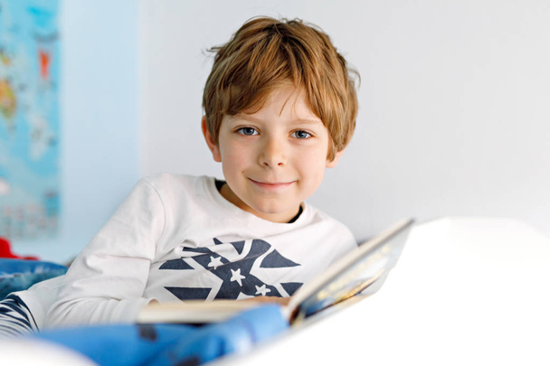 Cute blond little kid boy in pajamas reading book in his bedroom - Photo, Image