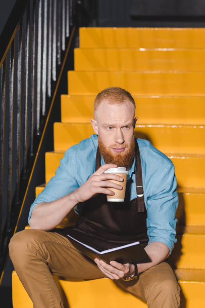 joven macho barista en delantal beber café de papel taza y celebración de libro de texto
 - Foto, imagen