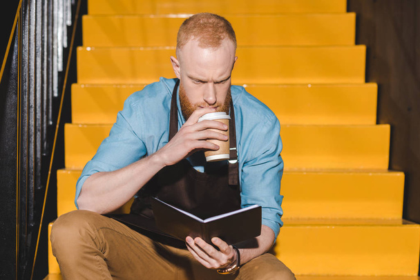 young male barista in apron drinking coffee from paper cup and looking on textbook  - Photo, Image