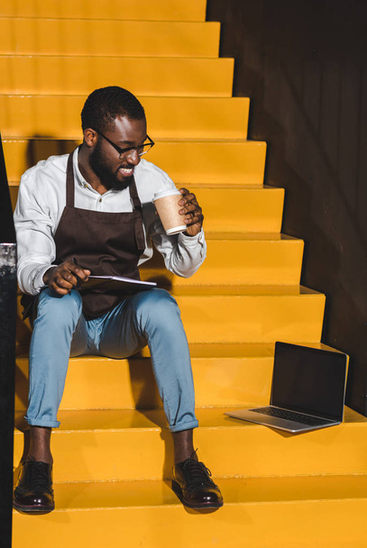 mosolyogva afroamerikai férfi barista tankönyv kávét iszik, és ült a lépcsőn, laptop - Fotó, kép