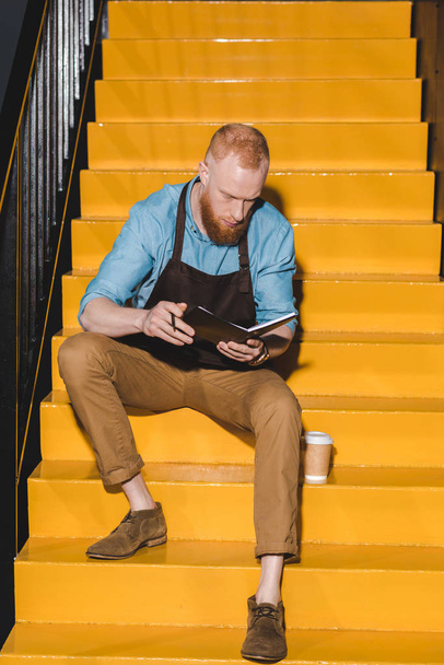 young male barista in apron holding textbook and sitting on stairs with disposable cup of coffee  - Photo, Image