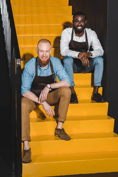 two smiling multiethnic owners of coffee shop in aprons sitting on stairs - 写真・画像