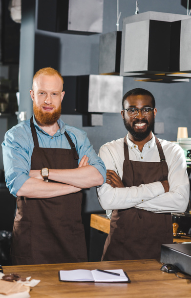 portrait of young multicultural owners of coffee shop in aprons standing with crossed arms in coffee shop - Foto, imagen