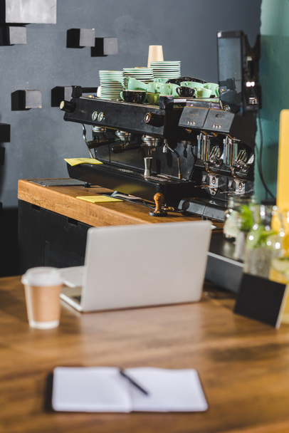 sisätilojen moderni kahvila kahvinkeitin, kannettava tietokone, paperi kuppi kahvia ja oppikirja pöydällä
 - Valokuva, kuva