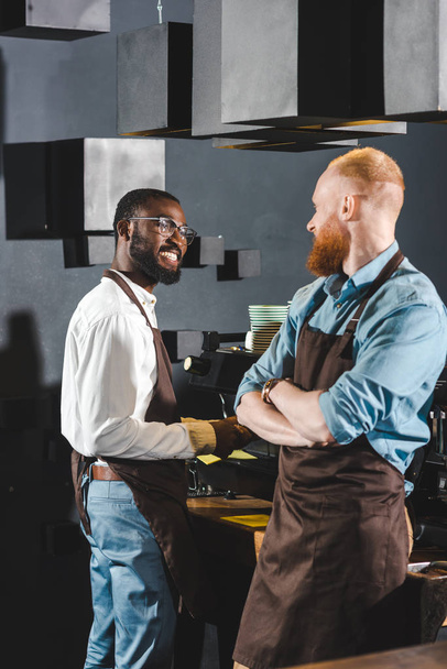 young smiling multiethnic owners of coffee shop in aprons talking in coffee shop - Foto, Bild